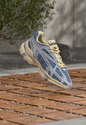 Blå og cremefarvet Lacoste-sneaker med mønstret mesh svæver over en træblok udendørs med sløret grøn baggrund og beton