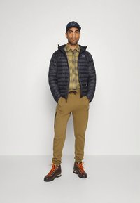 Casaco acolchoado preto, camisa xadrez verde, calças bege com bolsos laterais e botas castanhas com atacadores laranja. Modelo em pé, direito, contra um fundo liso.