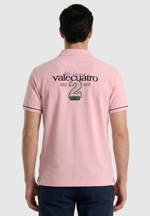 Hombre con una camiseta tipo polo rosa con texto azul marino y el número dos bordado en la espalda, de pie frente a un fondo liso.