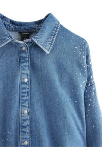 Camicia in denim di colore azzurro chiaro con colletto, bottoni a clip argentati e decorazioni sparse sulla spalla. Tessuto testurizzato con finitura sbiadita.