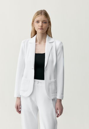 Junge Frau mit glatten blonden Haaren, die einen weißen Blazer und eine weiße Hose über einem schwarzen Oberteil trägt, steht vor einem einfarbigen hellen Hintergrund.