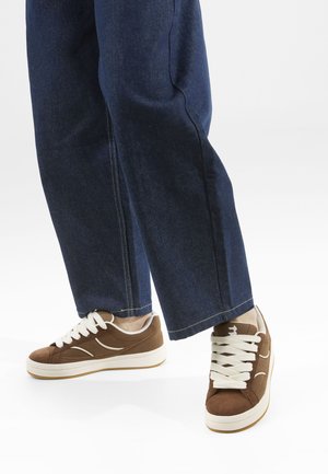 Baskets en cuir suédé marron avec des lacets blancs, contrastées par une semelle en caoutchouc blanche. Pantalon en denim bleu ample avec des coutures foncées et une coupe droite.