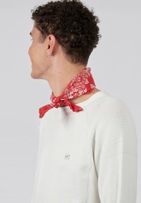 Bandana rouge avec motif paisley blanc noué autour du cou, porté sur un pull blanc avec des manches texturées et un petit logo.