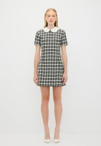 Schwarz-weißes Hahnentritt-Kleid mit tailliertem Design, kurzen Ärmeln und einem weißen, gewellten Kragen, kombiniert mit weißen spitzen Absatzschuhen.