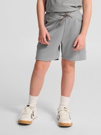 Kind draagt lichtgrijze short met trekkoord, witte sokken en witte sneakers met rubberen zolen, staand op een grijze ondergrond.