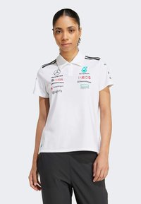 Hvid polo skjorte med knapkrave, sorte striber på skuldrene og flere sponsorlogoer trykt i forskellige farver. Subtil tekstur.