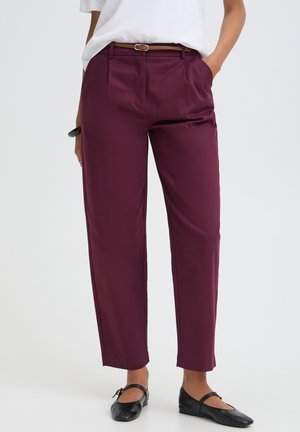 Personne portant un pantalon bordeaux taille haute avec une ceinture marron, une chemise blanche rentrée et des chaussures plates noires, debout, les mains partiellement dans les poches.
