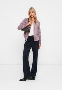 Jeune femme debout, portant une chemise à carreaux violette, un haut blanc, un pantalon large noir et des chaussures noires, tout en tenant un sac à bandoulière noir.