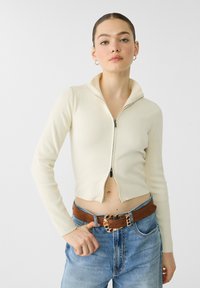 Pull court zippé crème avec texture côtelée, col montant et double zip. Associé à un jean en denim bleu taille haute et une ceinture marron.