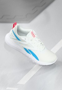 Scarpa sportiva Reebok bianca con dettagli blu, linguetta rossa sul tallone e lacci bianchi, posata su una superficie grigia chiara con linee bianche.