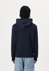 Hooded sweatshirt in navy, soft texture, kangaroo pocket, ribbed cuffs and hem, with a drawstring on the hood. Worn with light blue jeans.