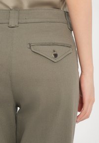 Gros plan sur la poche arrière d’un pantalon vert olive avec rabat boutonné, passant de ceinture, et une partie de la main d’une personne reposant à côté de la hanche.