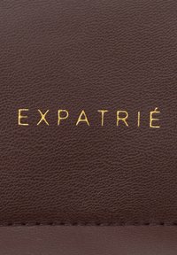 Brown leather with a textured surface, featuring embossed gold "EXPATRIÉ" lettering. Includes a stitched edge detail.