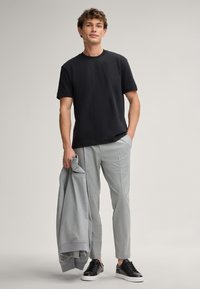 Schwarzes Kurzarm-T-Shirt kombiniert mit hellgrauen karierten Hosen und einer grauen Jacke, die in einer Hand gehalten wird. Schwarze Sneaker runden den Look ab.