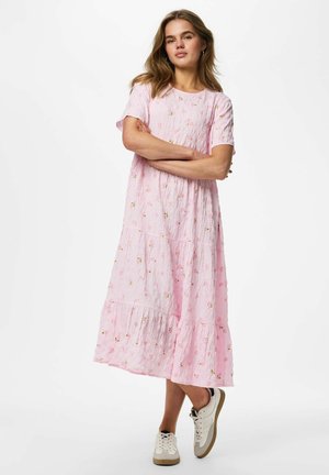 Kvinde står med armene over kors, iført en løstsiddende, lyserød blomstret midi-kjole og hvide sneakers, mod en ensfarvet hvid baggrund.