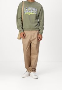 Oliwkowa bluza z białym i niebieskim logo "LACOSTE", połączona z beżowymi spodniami i białymi tenisówkami, trzymana z brązową torbą na ramię.