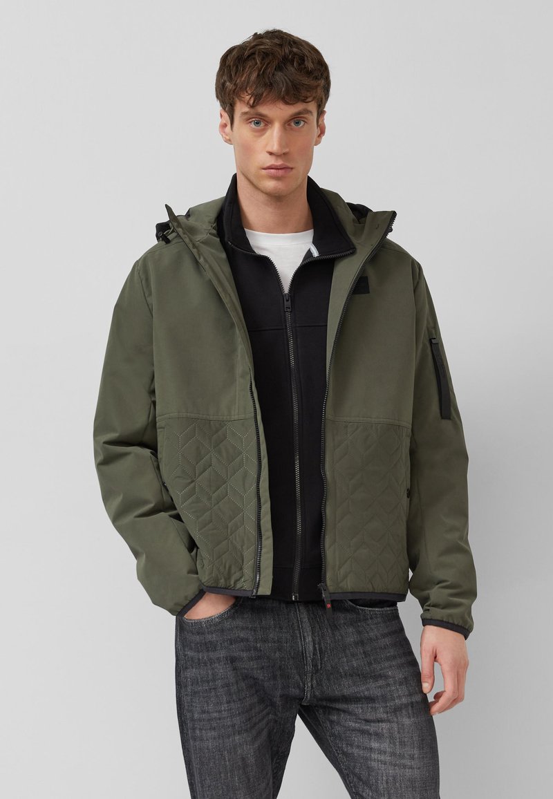 Jauns vīrietis valkā kapuci ar olīvu zaļas krāsas jaku ar šūtiem apakšējiem paneļiem, zem kā ir melna rāvējslēdzēja džemperis un tumši pelēnas džinsu bikses, stāvot pret pelēku fonu.