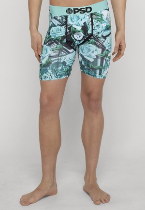 Mann iført turkis og svarte bokserunderbukser med blå rosemønster og sedler, stående barbeint mot ensfarget bakgrunn.