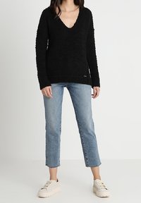 Pull noir texturé avec un col en V, associé à un jean court bleu clair et des baskets beiges. Tenue simple et décontractée sans logos visibles.