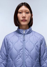 Lichtpaars gewatteerd jack met een hoge kraag, voorzien van afgeronde golfpatronen en een rits aan de voorkant. Textuur oppervlak en minimale brandingdetails.