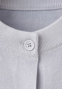 Svetlosivá pletená tkanina s rebrovaným golierom a jedným bielym štvordierkovým gombíkom zapnutým pri výstrihu.