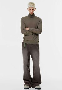 Olivgrüner Rollkragenpullover, weiche Textur. Breite braune Cordhosen mit flachem Vorderteil und einem grünen Akzentband. Graue Slipper mit strukturiertem Profil.