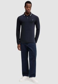 Granatowe polo z długim rękawem i paskowanym kołnierzykiem, z małym logo na piersi, w zestawie z ciemnymi jeansami i białymi trampkami.
