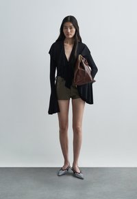 Black top with ruffled design, olive green shorts, and gray pointed flats. Brown leather bag held with a gold zipper accent. Minimalist style.