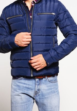 Man draagt een blauw gewatteerd jack met ritssluiting, dat hij aan het vastmaken is, gecombineerd met lichtblauwe spijkerbroek en een donker geruit overhemd eronder.