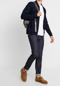 Man som går med marinblå jacka, vit t-shirt, mörka uppvikta jeans, bruna mockaskor och bär en grå ryggsäck på axeln.