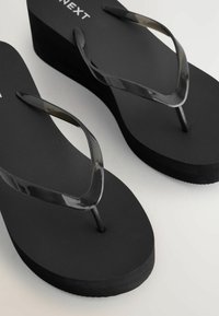 Sorte platforms-flipflops med teksturerede fodsenge og glansfulde sorte Y-formede remme på en lys baggrund.