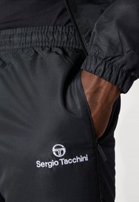 Čierna športová bunda s elastickým pásom, zipsovými vreckami a bielym vyšitým logom na ľavej strane. Štruktúrovaná tkanina.