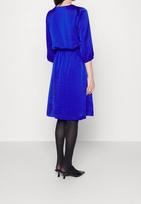 Vestido azul royal con mangas de tres cuartos, cintura ceñida, falda fluida y textura suave. Llevado con medias negras transparentes y zapatos de tacón mules.