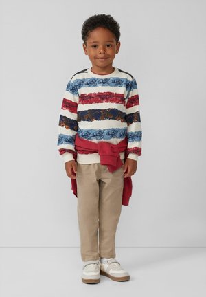 Gestreifter, mehrfarbiger Pullover in Blau, Rot und Weiß mit floralem Muster, kombiniert mit beigen Hosen und weißen Schuhen. Roter Pullover als Schal gebunden.
