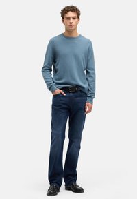 Jasnoniebieski dzianinowy sweter z okrągłym dekoltem, zestawiony z ciemnoniebieskimi dżinsami. Model nosi czarne eleganckie buty i czarny pasek. Gładkie tło.