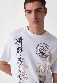 Vit bomulls-T-shirt med blandade grafiska motiv, inklusive text i svart och brunt, frimärken och blommönster. Rund halsringning, korta ärmar.