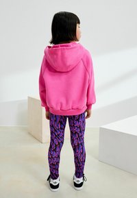 Rosa, huva sweatshirt, mönstrade leggings i lila och rosa, svarta sneakers med vita detaljer, barn som vänder ryggen mot kameran, minimalistisch bakgrund.