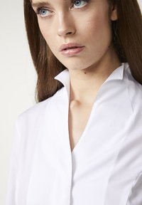 Weiße Bluse mit Kragen, die eine schlanke Passform, glatte Textur und ausgeprägte Nähte entlang des Kragens und der Vorderseite für einen modernen Look aufweist.