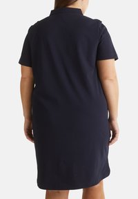 Femme portant une robe bleu marine foncé, courte aux genoux, à manches courtes, debout de dos face à la caméra sur un fond blanc.