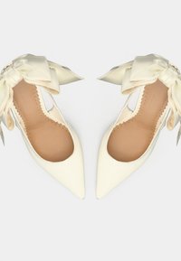 Talons aiguilles blancs en cuir verni avec bouts pointus, côtés ouverts et nœuds en satin à l'arrière. Texture lisse et semelles intérieure marron clair.