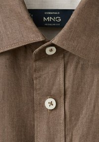 Camisa de lino marrón con cuello puntiagudo y dos botones blancos, etiqueta que dice "Essentials MNG Regular Fit" en el interior del cuello.