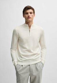 White pullover sweater with a half-zip collar, made from a smooth material. Features long sleeves and a fitted design. Paired with light gray trousers.