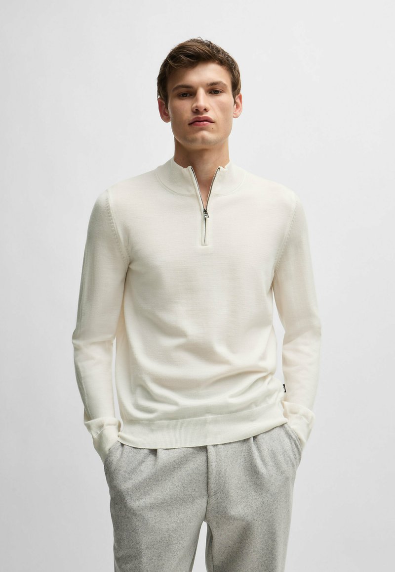 White pullover sweater with a half-zip collar, made from a smooth material. Features long sleeves and a fitted design. Paired with light gray trousers.