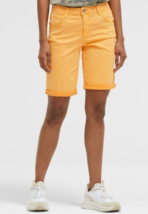 Person, die orange knielange Shorts mit umgeschlagenem Saum und weißen Sneakern trägt, vor einfachem Hintergrund stehend.