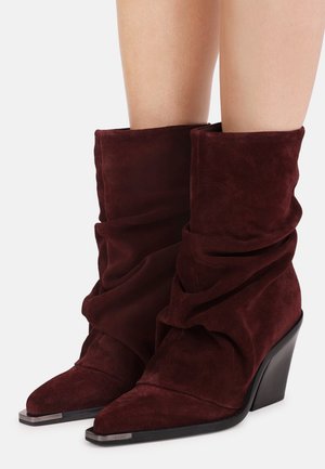 Bottines larges en daim rouge foncé avec bouts pointus, embouts métalliques et talons compensés hauts inclinés portées sur jambes nues.