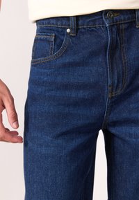 Un jean en denim bleu foncé présente une coupe droite avec des coutures visibles, une poche avant et une fermeture à bouton en métal.