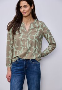 Femme portant un chemisier à manches longues à motifs verts et beiges avec des boutons, associé à un jean denim bleu, debout devant un fond clair uni.