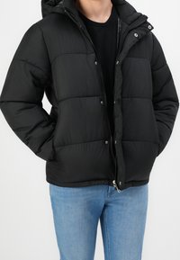 Chaqueta acolchada negra con capucha, cierre de cremallera y botones a presión. El material parece suave con una textura acolchada. Combinada con jeans claros.