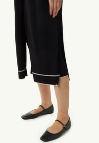 Schwarzes gestricktes Kleid mit weißen Akzenten am Saum und einem Seitenschlitz. Schwarze flache Schuhe mit einem schlichten Riemen.