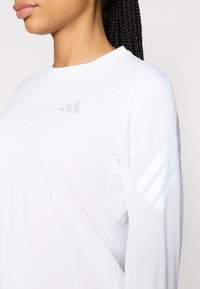 Camisa blanca de manga larga en tejido ligero con tres franjas diagonales de material reflectante en el hombro izquierdo.